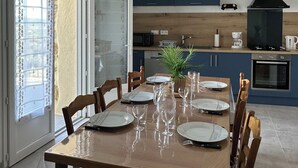 A dining area with a wooden table set for six, a kitchen in the background with modern appliances, and a sliding glass door leading to a balcony. - Gîte « Les Oliviers » Dans Mas Typiquement Ardéchois - 6 Personnes (Saint-Sauveur-de-Cruzières)