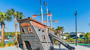 Een piratenspeelplaats met een glijbaan, omringd door palmbomen en een helderblauwe lucht.