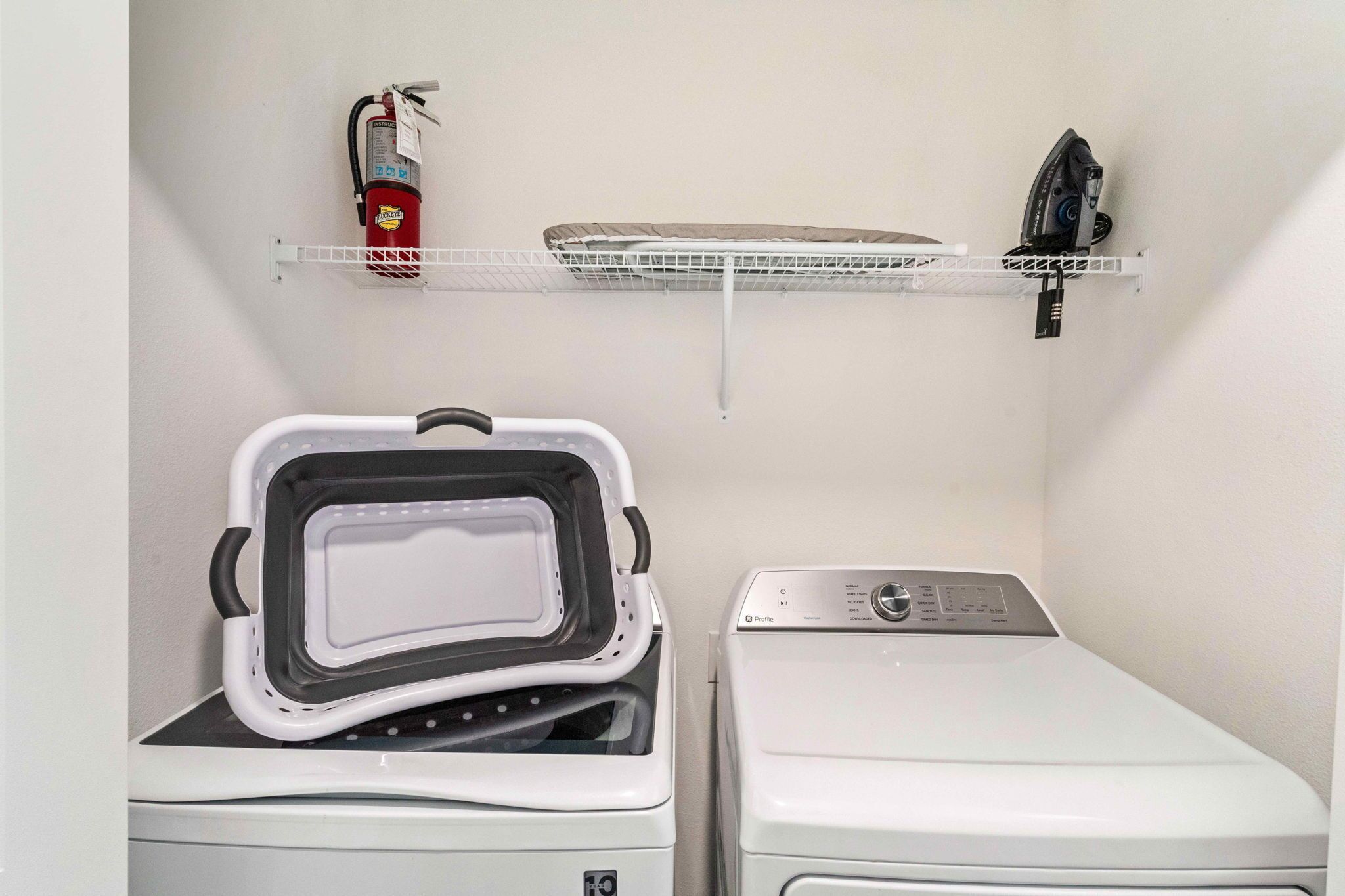A laundry area with a front-loading washer, a dryer, a shelf with a fire extinguisher, and an iron.