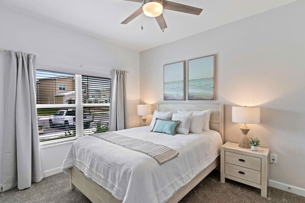 A bedroom with a bed, bedside lamps, a nightstand, a ceiling fan, and a window with curtains.