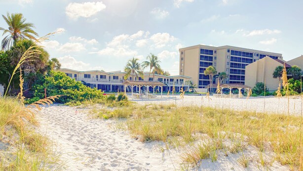 Ein Strandhotel mit einem Sandweg, der zu einem überdachten Bereich führt, Palmen und einem Gebäude mit vielen Fenstern.