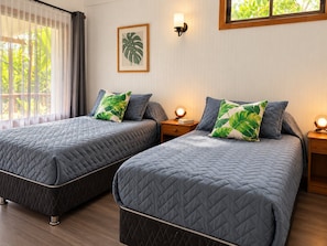 A hotel room with two beds, each with a quilted bedspread and a single pillow with a tropical leaf pattern. There is a framed plant on the wall, a bedside lamp, and a window with curtains.