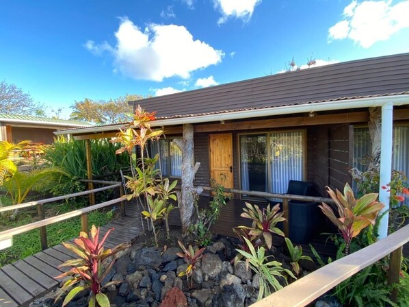 Une maison en bois dotée d’un porche, entourée de plantes tropicales et agrémentée d’une terrasse en bois.