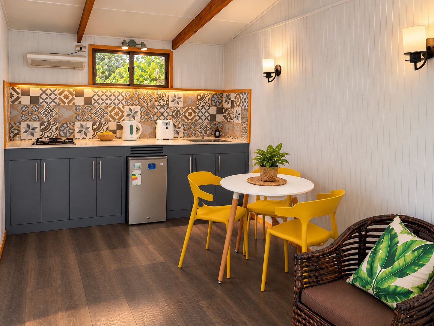 Une cuisine compacte avec un îlot central, un coin repas avec des chaises jaunes et un canapé en osier agrémenté d’un coussin à motif de feuilles vertes.