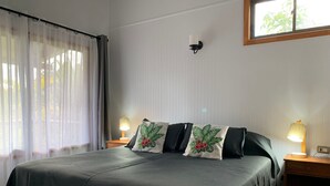 A neatly made bed with a dark green bedspread and two decorative pillows, a nightstand with a lamp, and a window with sheer curtains. - Comfortable and equipped cabin (Isla de Pascua)