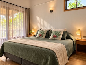 A bedroom with a bed, pillows, a nightstand, a lamp, a window with curtains, and a wall-mounted light fixture.