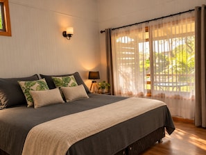 A bedroom with a large bed, a bedside lamp, a window with sheer curtains, and a view of greenery outside. - Comfortable and equipped cabin (Isla de Pascua)