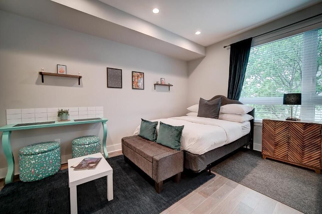 A modern bedroom with a bed, a desk, two ottomans, a small table, and a herringbone-patterned nightstand.