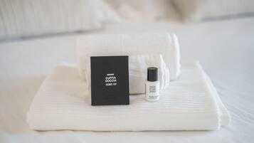 A neatly folded white towel, a black box labeled 'CUFFIA DOCCIA SHOWER CAP', and a small bottle of 'Oil for You' body lotion on a bed.