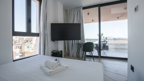 A hotel room with a large bed, a flat-screen TV, a small table with two chairs, and a view of the city through large windows. - Welcomely - Seamphony Suite vista Tavolara (Olbia)