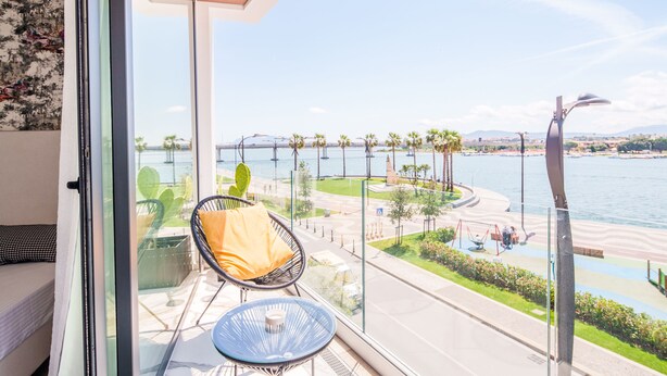 A balcony with a view of a waterfront promenade, a cactus plant, and a chair with a cushion. - Welcomely - Seamphony Superior Room (Olbia)