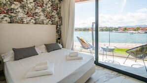 A hotel room with a large bed, two pillows, a neatly made bed with towels, a view of a body of water, and outdoor seating. - Welcomely - Seamphony Superior Room (Olbia)