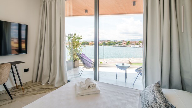 Une chambre d’hôtel dotée d’une grande fenêtre donnant sur un plan d’eau et sur une vue lointaine de la ville. On y trouve un lit avec une serviette soigneusement pliée, une chaise et une petite table.