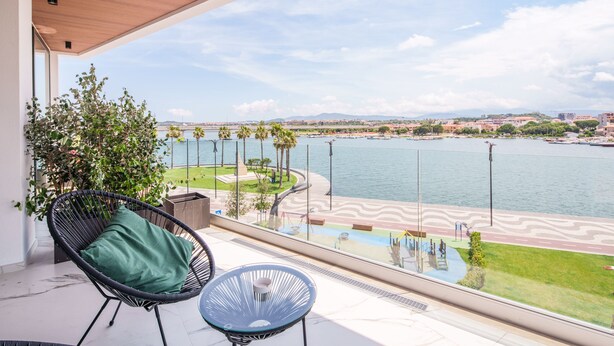 A balcony with a view of a waterfront promenade, a chair, and a small table. - Welcomely - Seamphony Deluxe Room (Olbia)