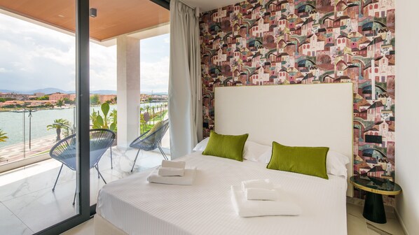 Une chambre d’hôtel avec un grand lit, deux oreillers verts, une tête de lit blanche et une vue sur un plan d’eau et des bâtiments.