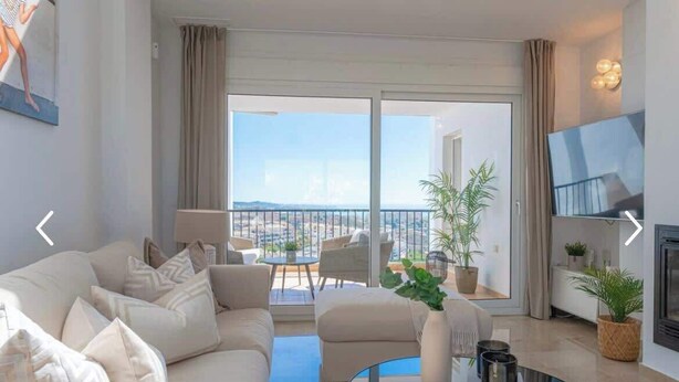 A modern living room with a sofa, coffee table, and a TV. There is a balcony with a view of the city.