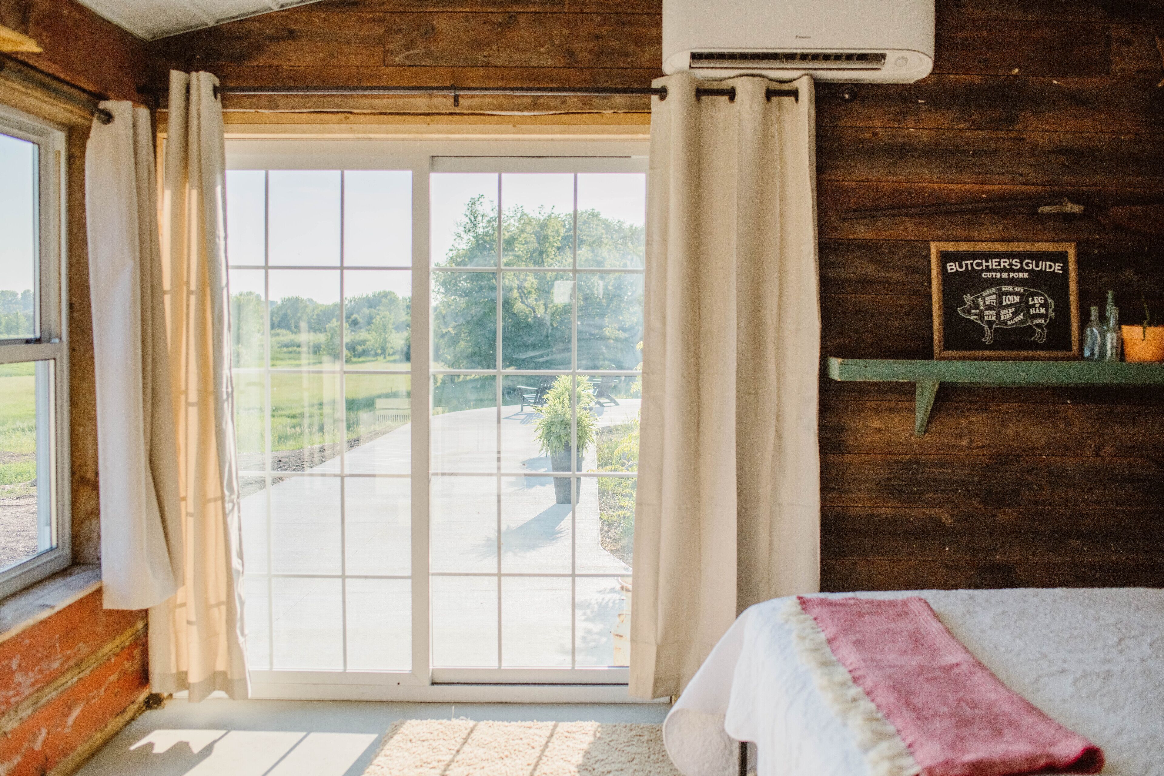 Una habitación con un amplio ventanal corredero a un jardín, una cama con manta blanca y rosa, y una pared de madera con un cartel de guía del carnicero.