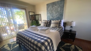 A bedroom with a checkered bedspread, a large painting above the bed, a nightstand with a lamp, and a view through a sliding door to an outdoor area.