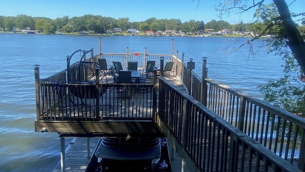 A wooden dock with chairs and a table, overlooking a calm lake with houses in the background.
