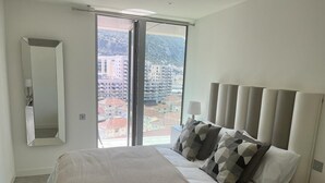 A modern hotel room with a large bed, a view of the city through a large window, and a minimalist design.