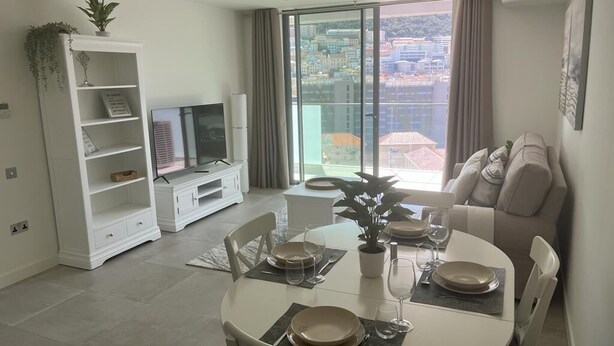 A modern living room with a dining area, a sofa, a TV, and a balcony with a city view.