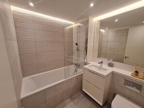 A modern bathroom with a glass-enclosed shower, a white bathtub, and a large mirror above the sink. - Eurocity Luxury Apartment (Gibraltar)