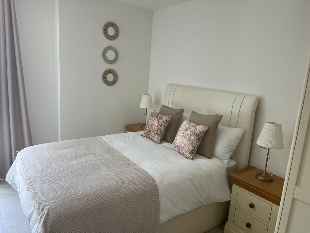 Una cama bien hecha con una colcha blanca y beige, dos almohadas y una mesita de noche con una lámpara.