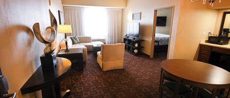 A hotel room with a sofa, armchair, dining table, and a TV.