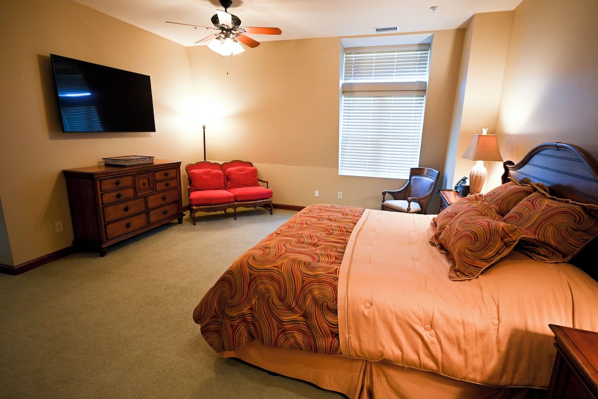 A bedroom with a bed, dresser, TV, ceiling fan, window, and chair.