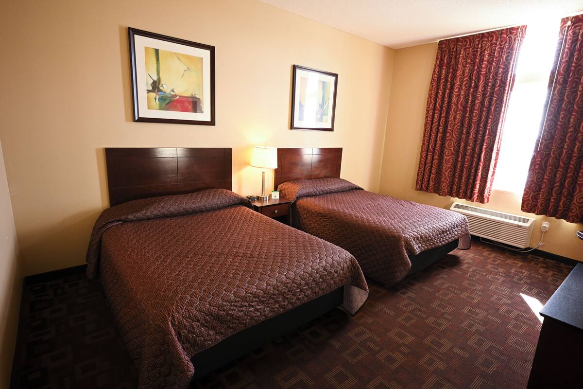 A hotel room with two beds, a nightstand, a lamp, a window with curtains, and a wall-mounted air conditioner.