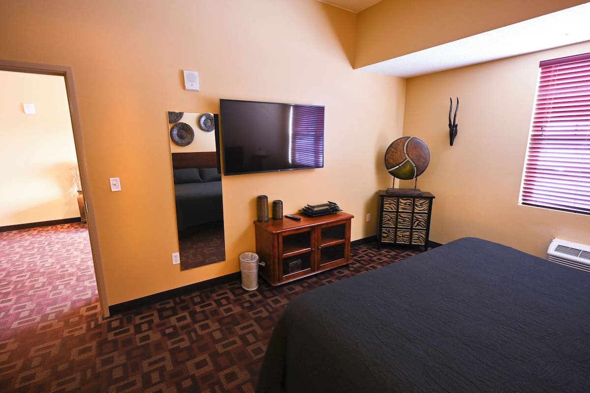 A hotel room with a large bed, a flat-screen TV mounted on the wall, a small wooden cabinet, and a decorative fan.