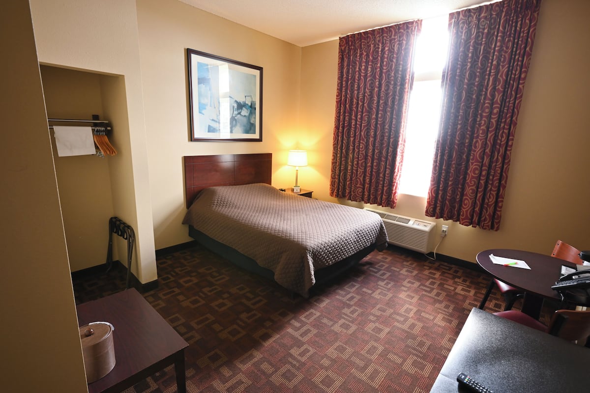 A hotel room with a bed, a desk, a chair, a table, a lamp, a painting on the wall, and a window with curtains.