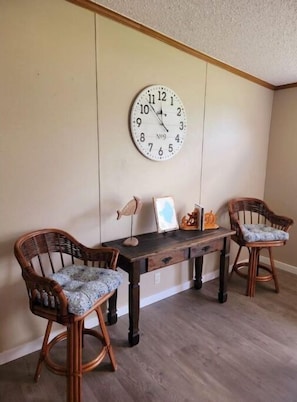 A dining area with a wooden table, two wicker chairs, a wall clock, and a small shelf with decorative items. - Mo's Lake "O" Retreat (Clewiston)