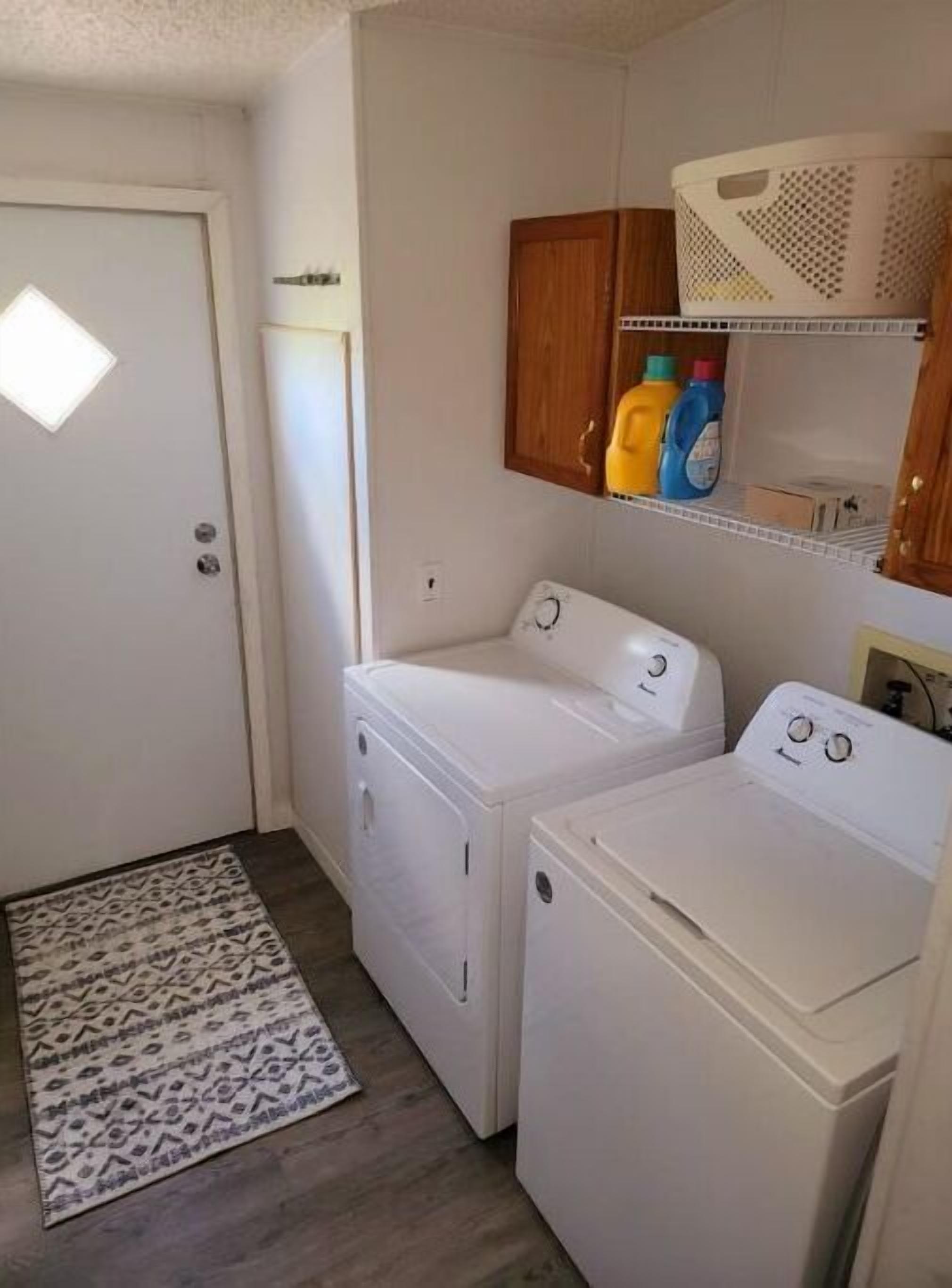 A laundry room with two white washing machines, a wooden cabinet above, and a patterned rug on the floor.