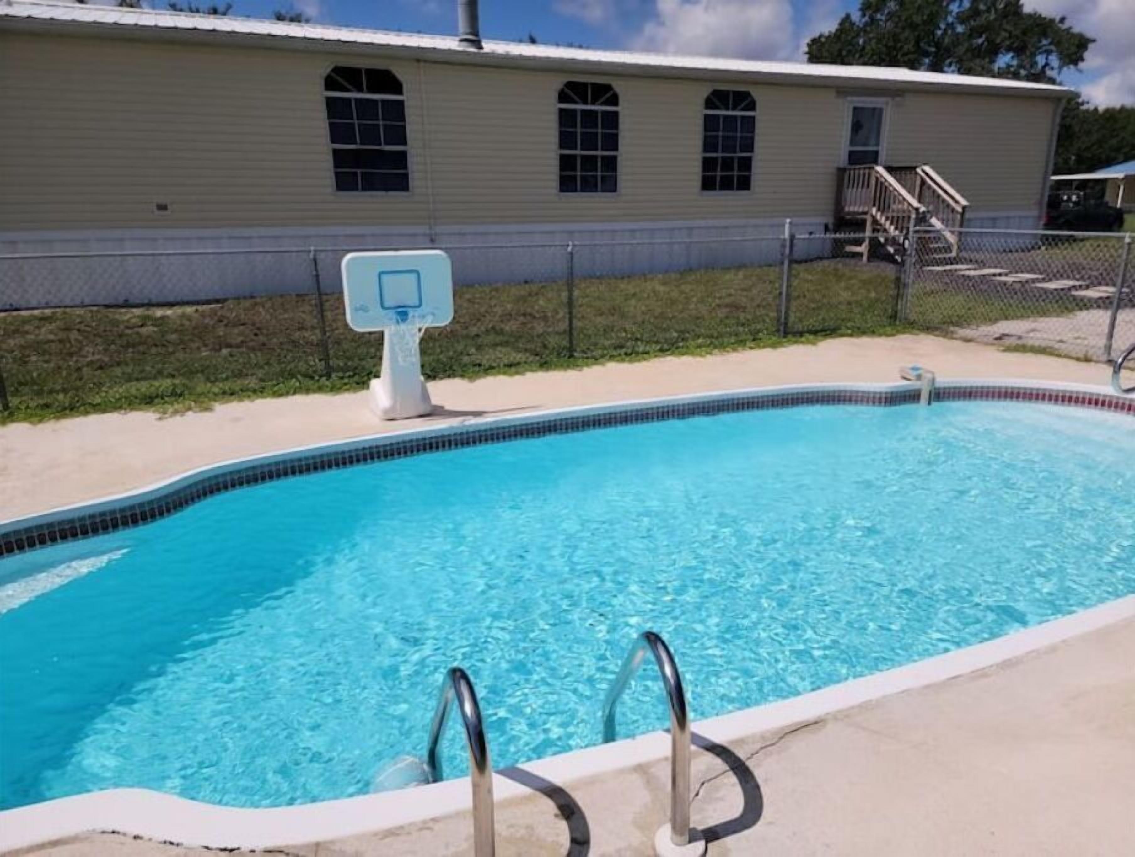 Una piscina con un aro de baloncesto, zona cercada y un edificio con ventanales arqueados al fondo.