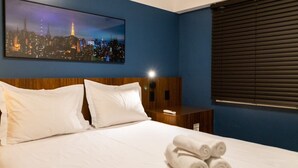A hotel room with a bed, white pillows, a nightstand, a remote control, and a cityscape painting on the wall. - Apto Peixoto Gomide (São Paulo)