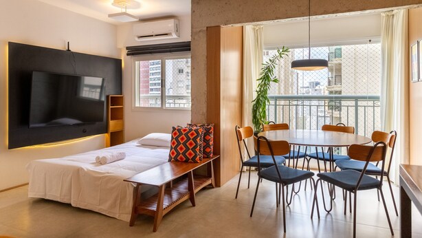 A modern living room with a flat-screen TV, a dining table with chairs, a sofa with patterned cushions, and a large window with a view of buildings. - Apto Peixoto Gomide (São Paulo)