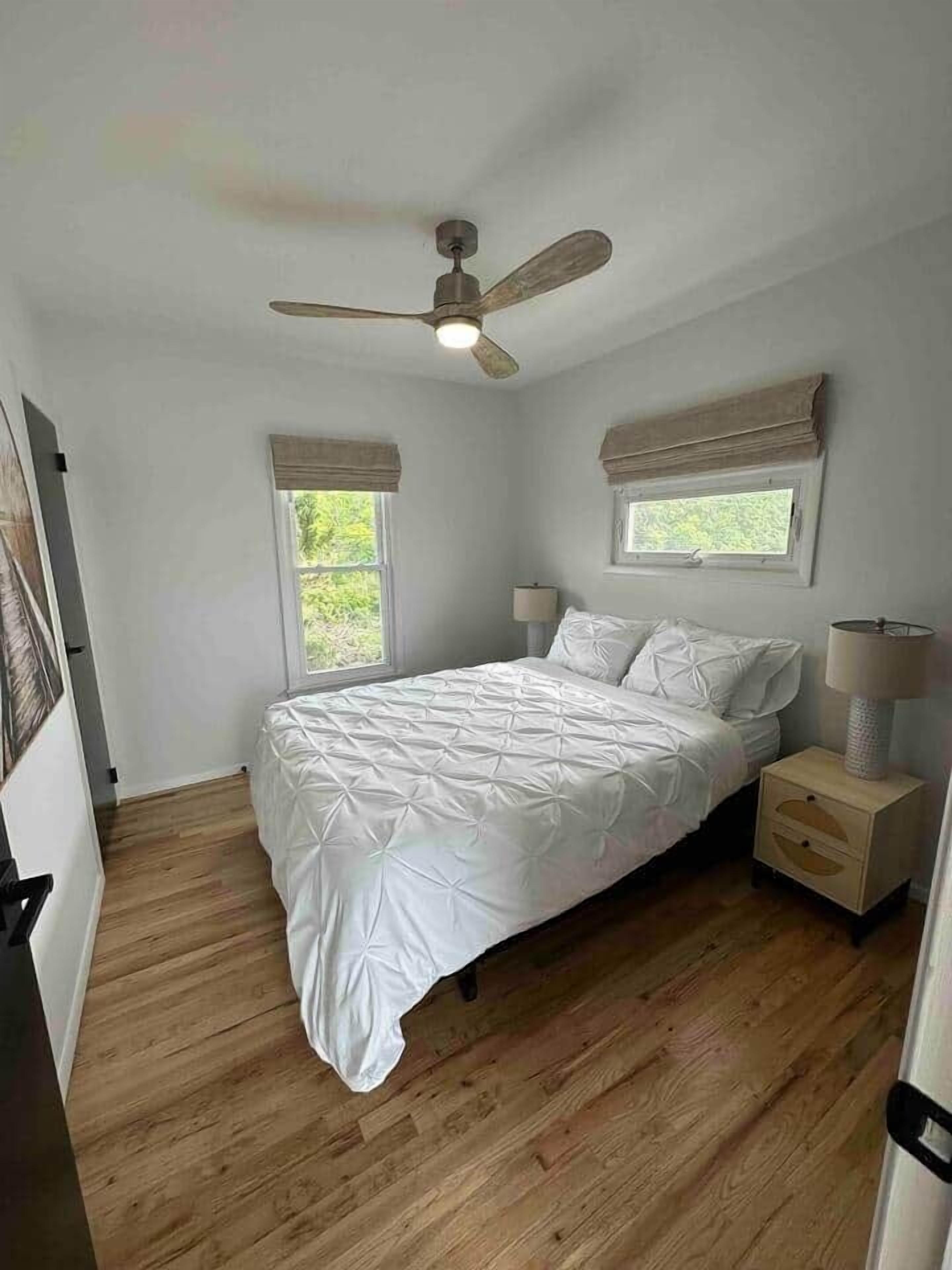 Ein Schlafzimmer mit Bett, Deckenventilator, Fenster mit Vorhängen, Nachttisch und Lampe.