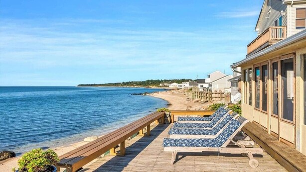 A wooden deck with lounge chairs and a potted plant, overlooking a beach and ocean.