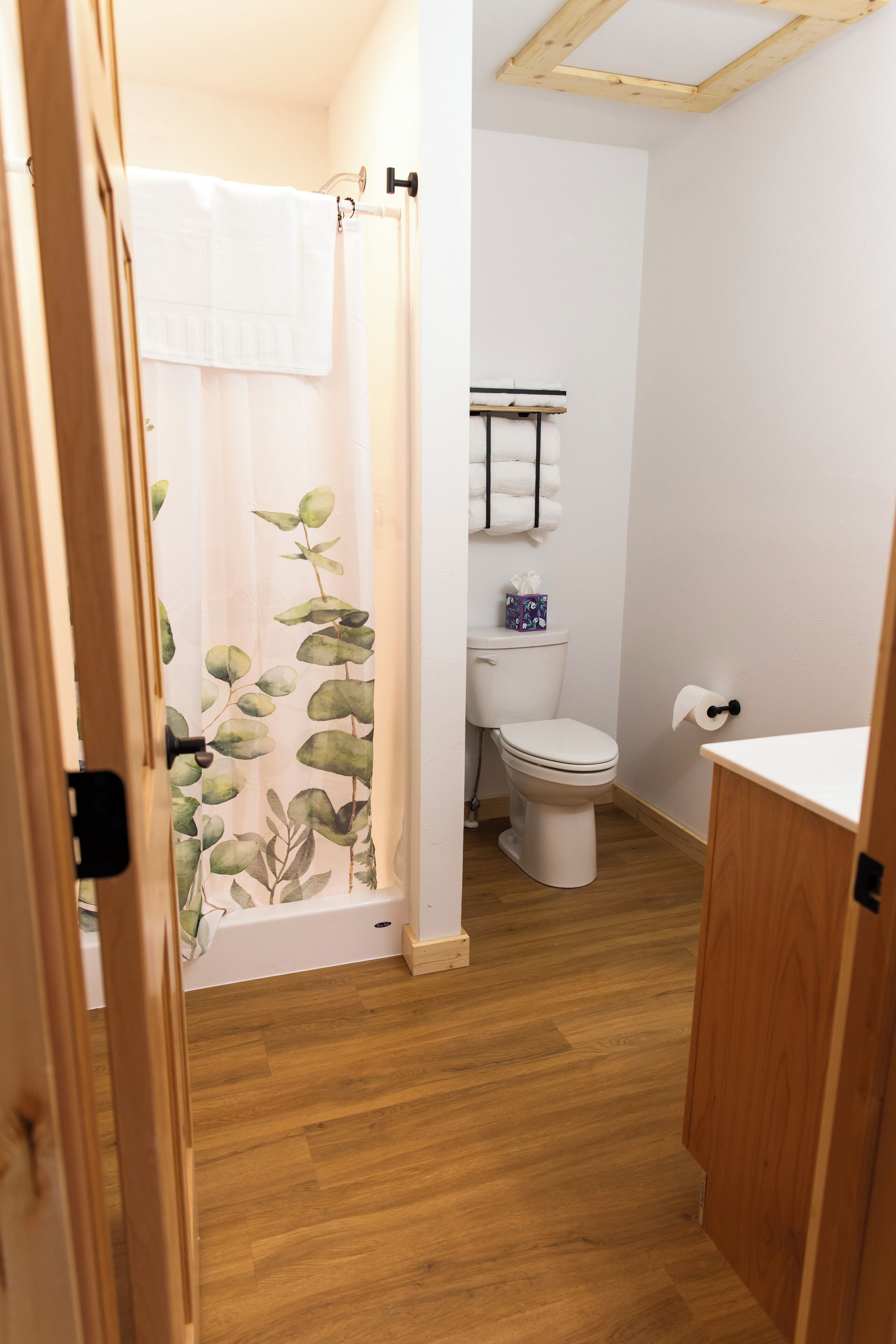 A bathroom with a toilet, a shower curtain with a leaf pattern, and a wooden floor.