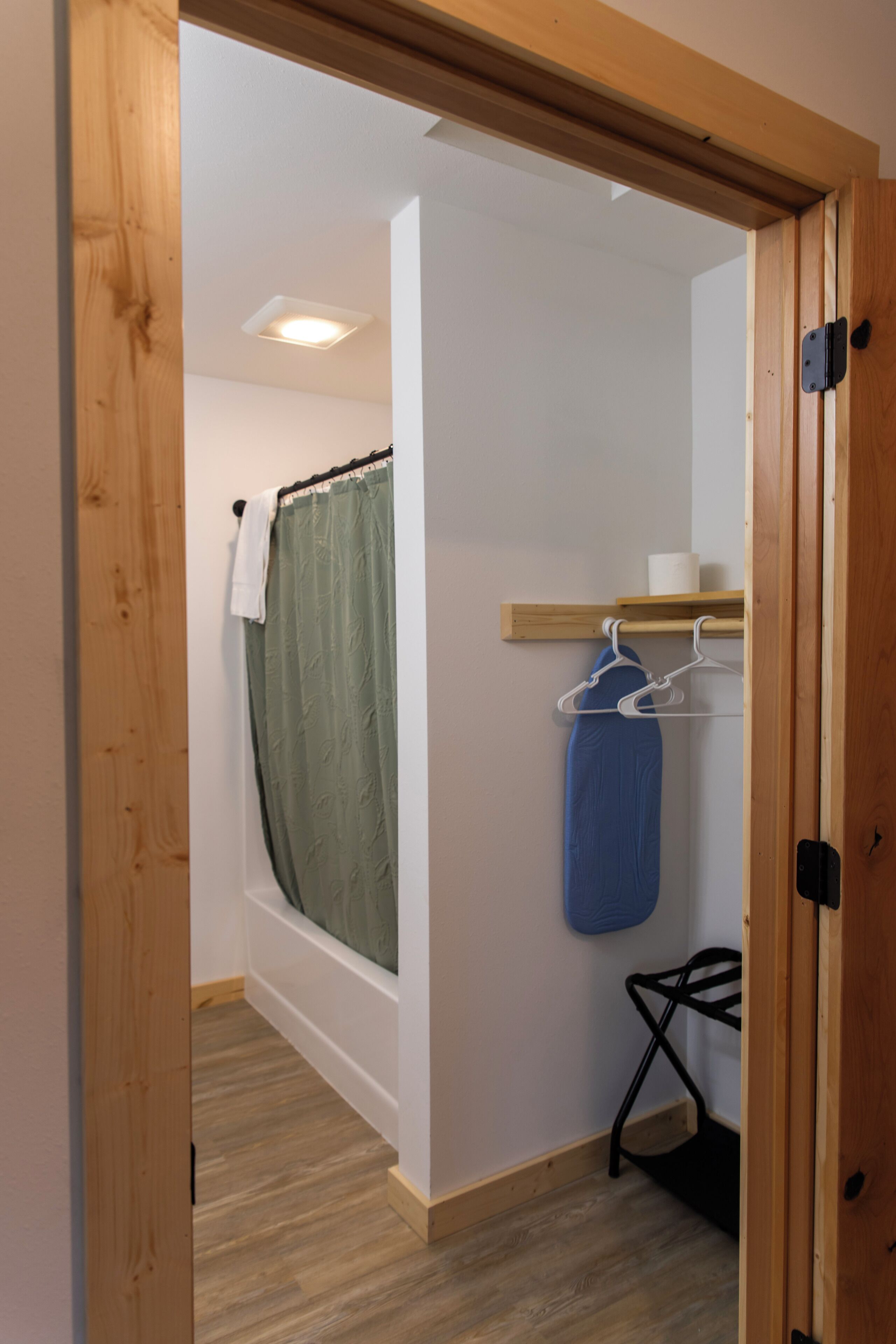 A narrow hallway with a wooden door frame, a white wall, a wooden floor, a small shelf with a white towel, a blue hanging bag, and a black chair.
