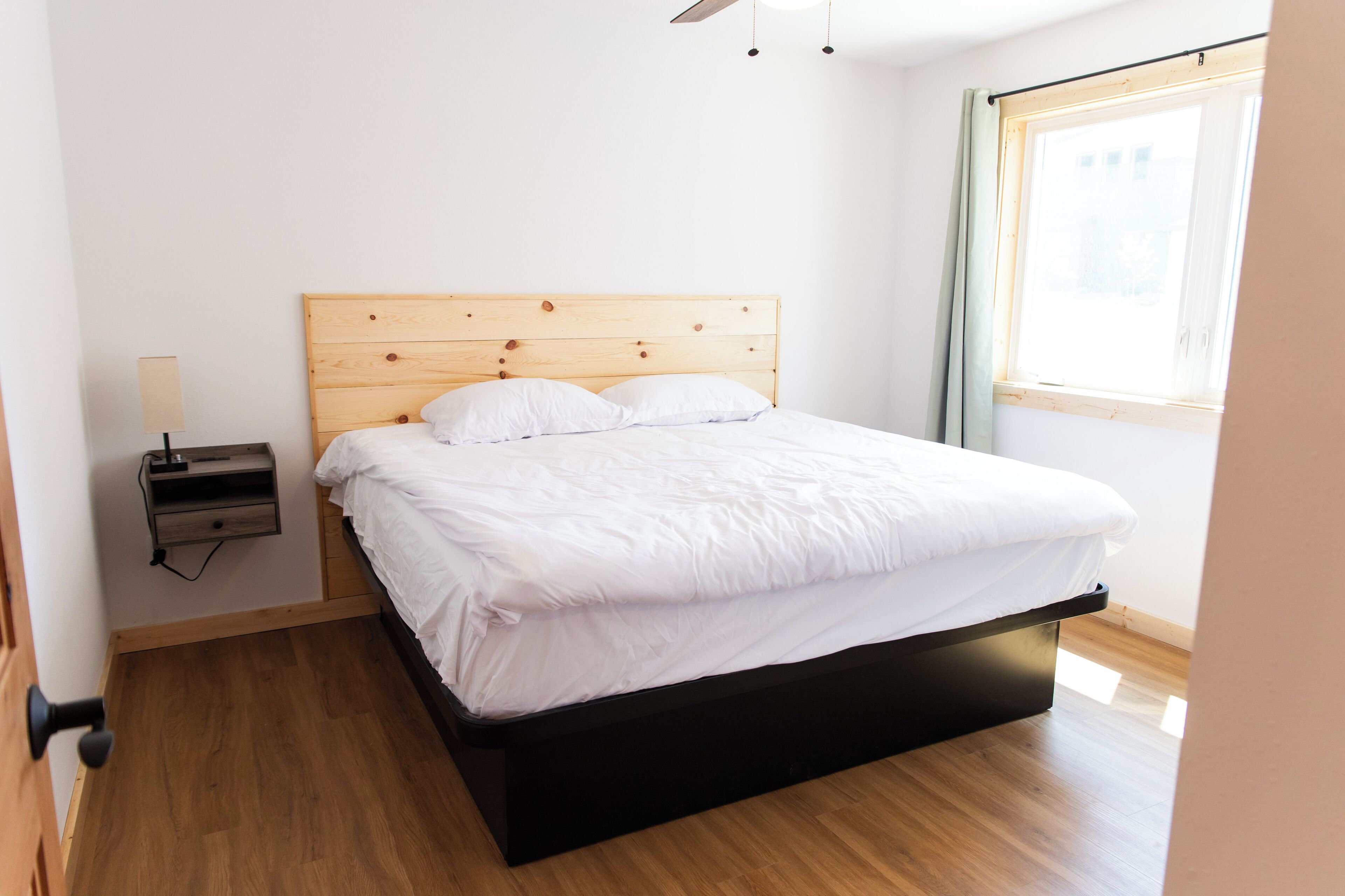 A neatly made bed with a wooden headboard, a bedside table with a lamp, and a window with a view of the outside.