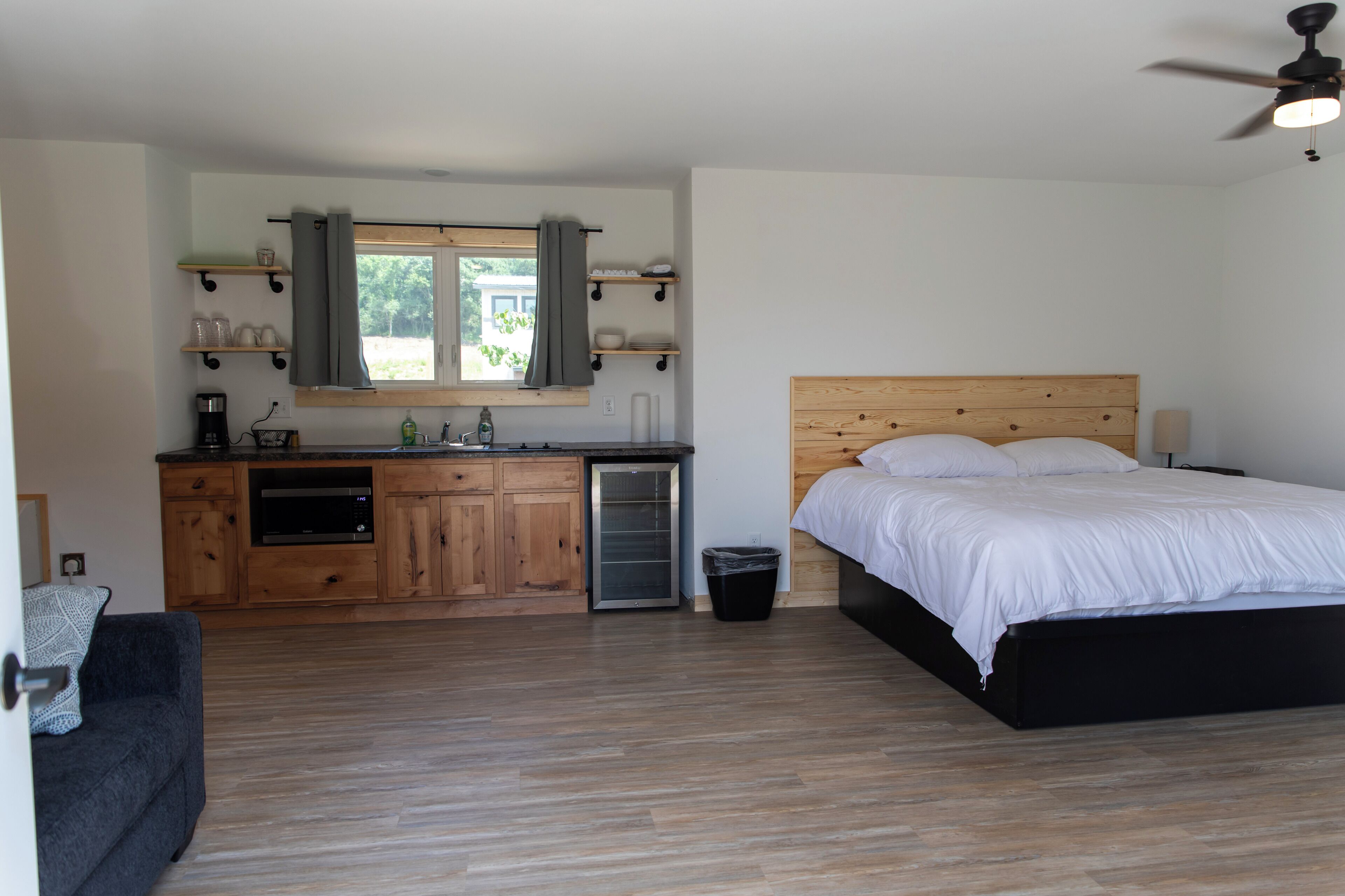 A bedroom with a wooden headboard, a bed with white bedding, a small kitchenette with a microwave, and a ceiling fan.