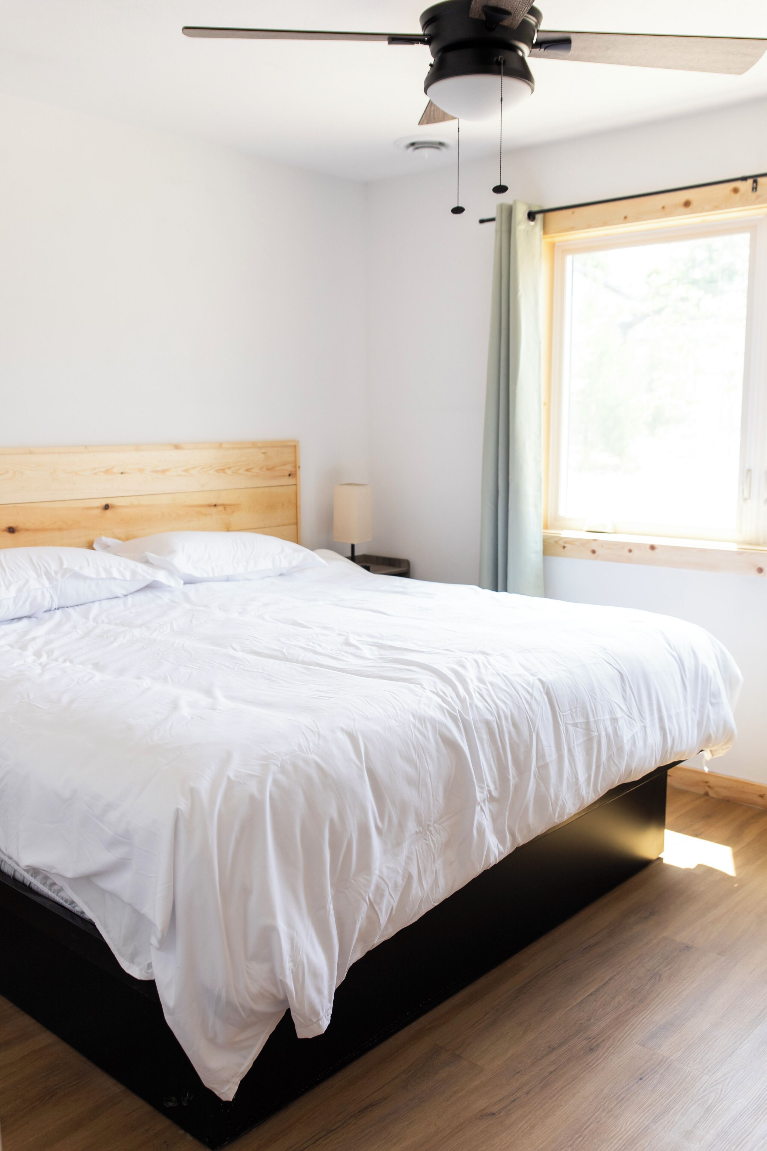 A neatly made bed with a wooden headboard, a ceiling fan, a bedside lamp, and a window with curtains.