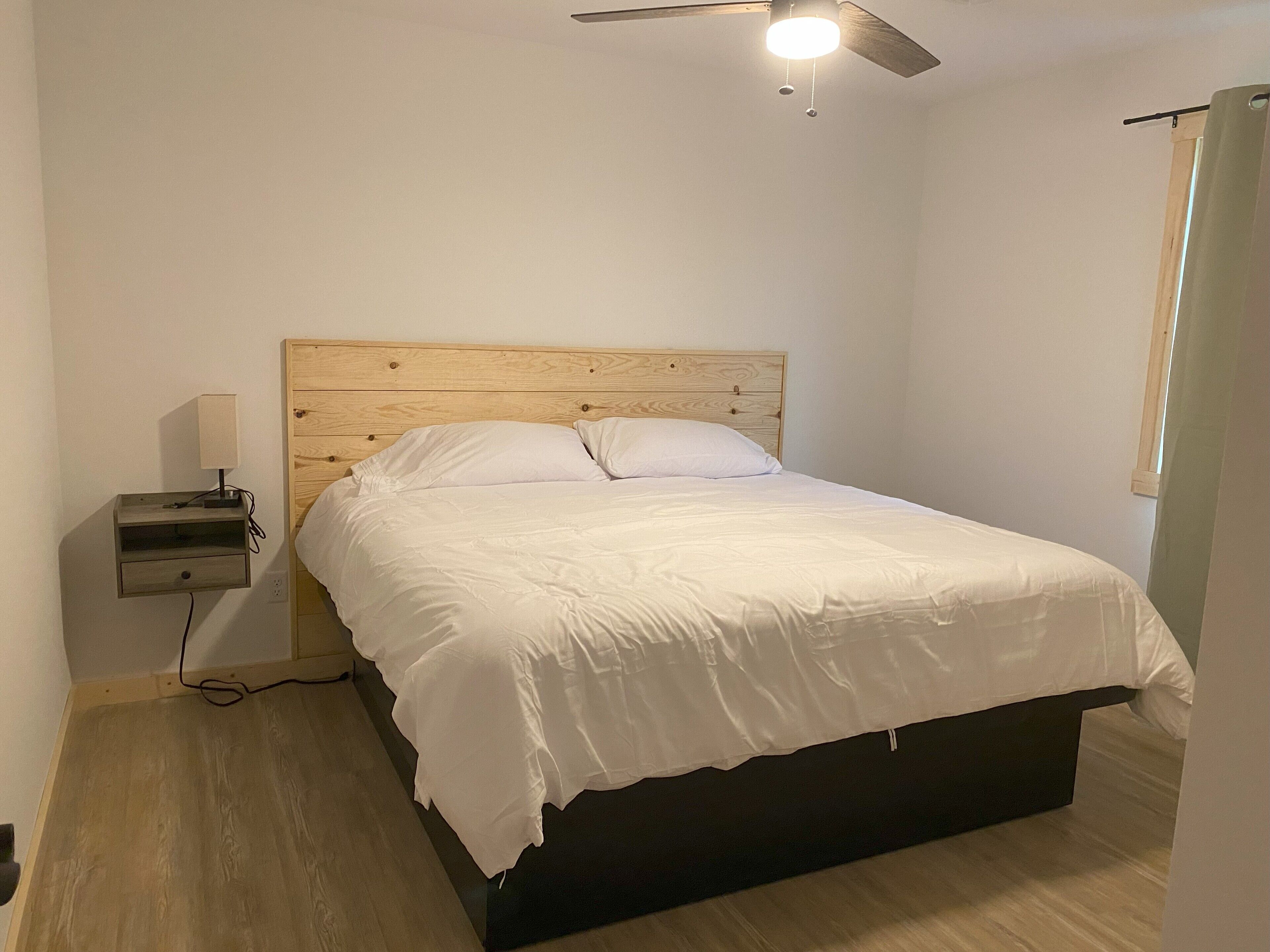 Un dormitorio con cabecera de madera, cama con ropa de cama blanca, mesita de noche con lámpara y ventilador de techo.
