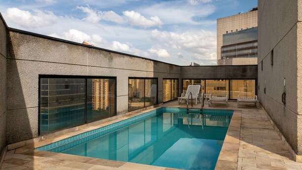 Ein Pool mit gefliesten Rändern, umgeben von einer Steinterrasse. Liegestühle und ein verglaster Bereich mit Blick auf ein Gebäude.