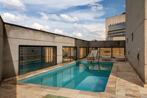 A swimming pool with tiled edges, surrounded by a stone patio. There are lounge chairs and a glass-enclosed area with a view of a building. - Apto Fidêncio Ramos 64 (São Paulo)
