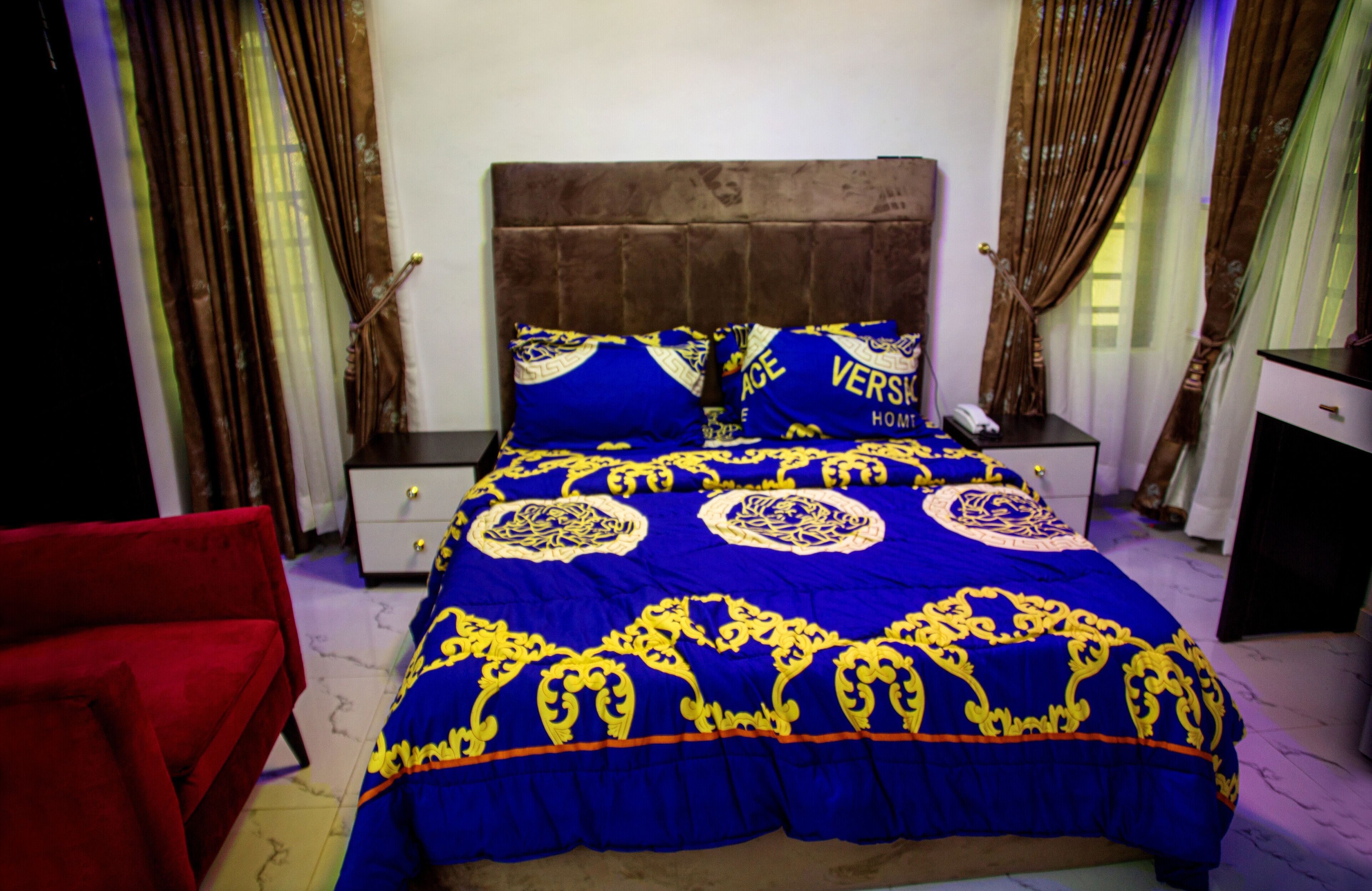 A bedroom with a large bed, a red chair, a nightstand with a phone, and curtains.