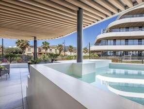 A modern pool area with a curved edge, a covered seating area, and a multi-story building in the background.