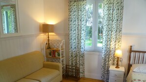 Quarto aconchegante com piso de madeira, sofá bege, mesinhas de cabeceira branca e pequena com uma luminária, cabeceira de madeira e uma janela com cortinas florais.